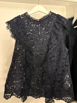 Mörkblå spetsblus med volang - Snygg mörkblå blus i spets med blommigt mönster och volangdetaljer längs axlarna. Blusen är ärmlös och har en rund halsringning med spetskant. Perfekt för dig som gillar romantiska detaljer och en feminin look.