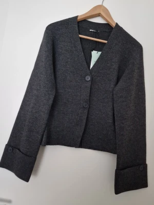 Mörkgrå kofta från Gina Tricot XS - Snygg mörkgrå kofta från Gina Tricot i storlek XS med två stora knappar framtill och breda uppvikta ärmslut. Koftan har en rak, något kortare passform och V-ringning. Perfekt att slänga över en t-shirt eller topp för en chill look. Ny med prislapp