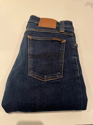 Nudie Jeans - Snygga mörkblå jeans från Nudie Jeans med klassisk femficksmodell och kontrastsömmar. Jeansen har en rak passform och är tillverkade i denim med en liten läderpatch bak i midjan. Perfekta för en avslappnad och stilren look.