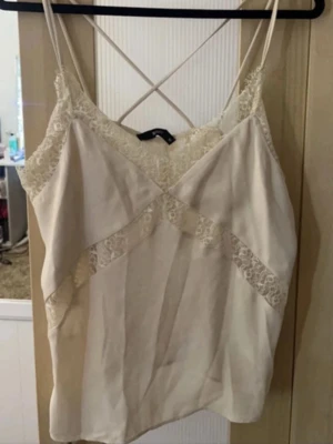 Beige linne med spetsdetaljer - Sött beige linne med tunna axelband och v-ringning. Dekorerat med spets längs halsringningen och diagonalt över framsidan. Lätt och luftigt material, perfekt för sommaren 