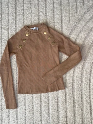 Zara tröja S kamelfärgad beige/brun guldknappar ribbad stickad topp med knappar - Strl: S. Helt ny och oanvänd. Snygg beige/brun ribbad stickad topp från Zara med lång ärm och dekorativa guldfärgade knappar på axlarna. Perfekt till jeans eller kjol för en trendig look.