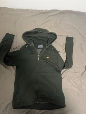 Mörkgrön zip hoodie från Lyle & Scott - Mörkgrön hoodie med dragkedja från Lyle & Scott. Klassisk huva med snören och gul logga på bröstet. Två fickor framtill och ribbade muddar vid ärmslut och nederkant. Perfekt för en avslappnad och stilren look.