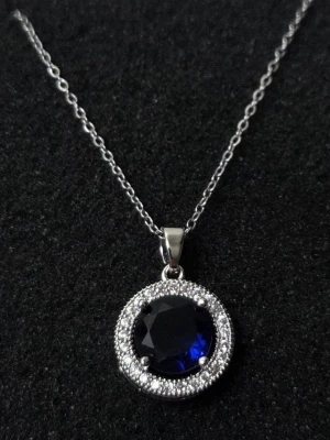 Silverhalsband med blå sten och pärlor i rostfritt stål  - Snyggt halsband i silverfärg med en rund, mörkblå sten i mitten omgiven av små glittrande pärlor. Kedjan är tunn och elegant, och hänget har en klassisk, tidlös design som verkligen sticker ut. Perfekt för dig som gillar stilrena smycken med en touch av färg.