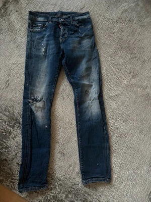 Dsquared Jeans - Säljer ett par blå jeans med slitningar och hål på knäna. Jeansen har en smal passform och detaljer som ljusa tvättade partier och små färgstänk. Perfekta för en avslappnad och trendig look. | Storlek 42 |