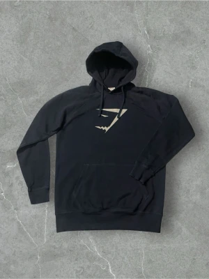 Mörkblå hoodie från Gymshark | M - Snygg mörkblå hoodie från Gymshark med stor vit logga på bröstet. Hoodien har huva med dragsko och en stor magficka. Tillverkad i mjukt material som passar perfekt till chill eller träning. Klassisk passform och stilren design.