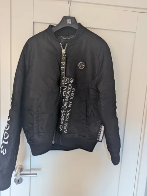 Svart bomberjacka Philipp Plein - Snygg svart bomberjacka från Philipp Plein med vita tryckta detaljer och logga på bröstet. Jackan har dragkedja framtill, ribbade muddar och coola textdetaljer längs dragkedjan och ärmen. Materialet är glansigt och ger en modern streetwear-look.