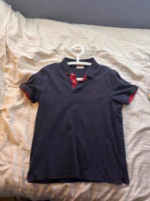 Marinblå piké från Gant med röda detaljer - Snygg marinblå pikétröja från Gant med röda detaljer vid knappar och ärmslut. Klassisk krage och korta ärmar. Tillverkad i mjuk bomull som känns skön mot huden. Perfekt för dig som gillar stilrena och sportiga plagg.