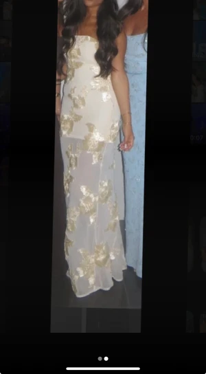 Bella barnett Klänning  - Bella barnett, Säljer en elegant långklänning i vit mesh med guldiga blommiga broderier. Klänningen är axelbandslös och har en figurnära passform som ger en snygg siluett. Perfekt för dig som vill sticka ut med en glamorös look.