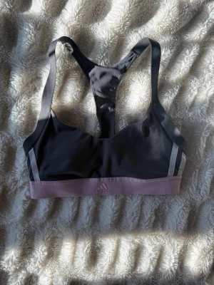 Svart sport-bh från Adidas - OBS: lappen med storlek är avklippt, men jag skulle tippa på att det är en storlek XS. Sport-bh från Adidas i grå med bred, ljuslila resår under bysten och grå detaljer längs axelbanden. Racerback-modell för bra stöd och rörelsefrihet. Tillverkad i ett stretchigt funktionsmaterial som passar perfekt till träning. 