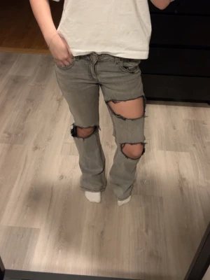 Grå slitna ripped jeans bootcut - Säljer ett par grå bootcut jeans med stora ripped hål över knäna och låren. Jeansen har fickor och hålen är hemmagjorda.