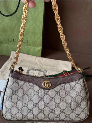 Gucci GG Supreme axelremsväska - Säljer en snygg Gucci axelremsväska i GG Supreme canvas med bruna läderdetaljer och guldfärgad kedja. Väskan har det klassiska GG-mönstret och en elegant form med dragkedja upptill. Perfekt för dig som gillar lyxiga accessoarer och tidlös design.