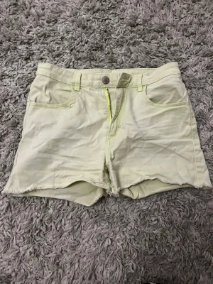 Ljusgröna/gula jeansshorts med rå kant - Snygga ljusgula shorts i jeansmaterial med råa kanter nertill. Klassisk femficksmodell med bälteshällor och knapp- samt dragkedjestängning framtill. Perfekta för varma dagar och enkel att matcha med olika toppar.