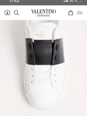 Valentino Garavani vit/svart sneaker - Stilren sneaker från Valentino Garavani i vitt läder med en bred svart läderdetalj över snörningen. Klassisk låg modell med subtila nitar på sulan och guldfärgad logga på plösen. Perfekt för dig som gillar exklusiv och modern design.