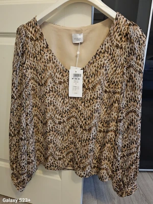 Mönstrad blus från Vila med v-ringning - Snygg blus från Vila med v-ringning och ett brunt och beige abstrakt mönster. Blusen har långa ärmar med lätt puff och resår vid ärmslut. Tillverkad i ett tunt, luftigt material som ger en skön känsla. Perfekt för dig som gillar trendiga mönster och avslappnad stil.