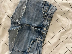  jeansshorts - Säljer ett par blå jeansshorts med råa, fransiga kanter och klassisk femficksdesign. Shortsen har bälteshällor, knapp och dragkedja framtill. Perfekta för sommaren och ger en avslappnad streetstyle-look.