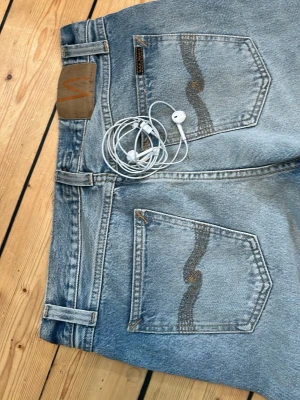 Ljusblå jeans från Nudie Jeans - Säljer ett par ljusblå jeans från Nudie Jeans med klassisk femficksmodell och snygga kontrastsömmar. Bakfickorna har den ikoniska vågiga sömmen och läderpatch bak i midjan. Perfekta för en avslappnad och trendig look. Ändast använda en gång. 