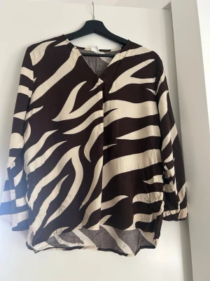 Zebramönstrad blus med V-ringning - Snygg blus med zebramönster i brunt och beige. Blusen har V-ringning och långa ärmar med avslappnad passform. Perfekt för dig som vill sticka ut med ett djurmönstrat plagg. Tunn och luftig, passar bra till både jeans och kjol.