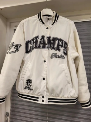 Vit bomberjacka CHAMPS från H&M S - Snygg vit bomberjacka från H&M med svart text 'CHAMPS Series' på bröstet och broderade detaljer som '93' på ärmen. Jackan har svarta och vita ränder på krage, ärmslut och nederkant samt tryckknappar framtill. Perfekt för en sportig och trendig look.
