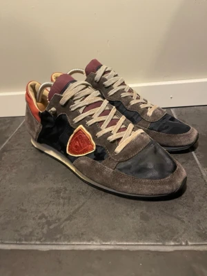 Philippe Model sneakers grå/svart - Snygga sneakers från Philippe Model i grå mocka och svart textil med vinröda och orange detaljer. Skorna har beige snören och en låg siluett. Klassisk logga på sidan och bekväm innersula. Perfekta för dig som gillar streetstyle och unika detaljer.