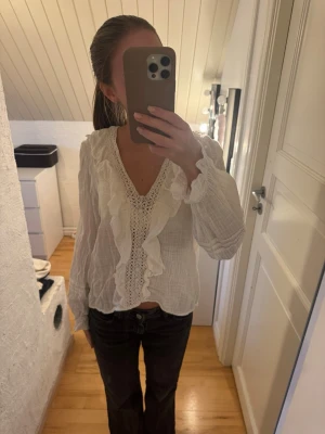 Vit blus med volanger och spets - Supersöt vit blus med långa ärmar, volangdetaljer längs v-ringningen och spets framtill. Blusen har en luftig och lätt struktur, perfekt för en trendig och romantisk look. Materialet känns mjukt och bekvämt mot huden.