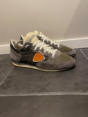 Philippe Model grå sneakers - Snygga sneakers från Philippe Model i grå mocka och mesh med vita detaljer och orange logotyp på sidan. Skorna har vita snören med tryck och en låg silhuett. Perfekta för dig som gillar streetwear och vill ha något unikt på fötterna.