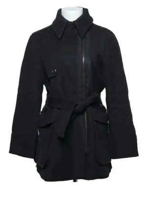Svart kappa med bälte - Stilren svart kappa med klassisk krage, knytskärp i midjan och stora fickor med lock. Den har dragkedja framtill, dekorativa knappar och slits bak. Kappan är i mycket bra skick och säljer då den är för liten för mig 