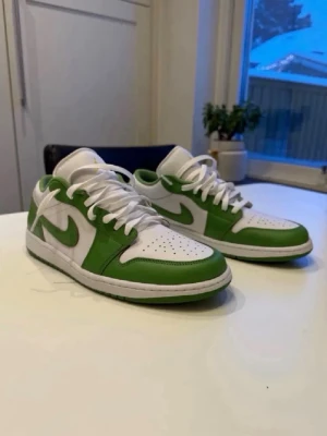Nike Air Jordan 1 Low - Säljer ett par Nike Air Jordan 1 Low sneakers i vitt och grönt. Skorna har klassisk låg siluett, vita skosnören och perforerad tåbox. Tillverkade i läder med gröna detaljer och ikonisk swoosh på sidan. Perfekta för dig som gillar streetwear och sneakerskultur. Helt ny aldrig använd köpte för 1349kr 
