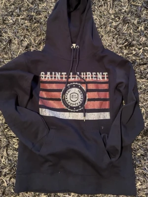 Blå hoodie från Saint Laurent - Blå hoodie från Saint Laurent med tryck framtill i rött, vitt och blå. Stor huva med dragsko och känguruficka. Snygg streetstil med Saint Laurent-logga och grafiskt mönster. Står S men sitter som XS. Passar till folk som är mellan 168-178. Priset kan diskuteras vid snabbaffär! Vid minsta lilla fundering skriv så löser vi något!