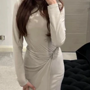 Beige långklänning från She's Out - Jättefin helt ny stilren beige långklänning från She's Out med lång ärm och snygg knutdetalj i midjan. Klänningen har en figurnära passform och är tillverkad i ett mjukt, stretchigt material som ger en elegant siluett. Prislapp kvar. Storlek onesize men skulle säga den ändå är xs-s.