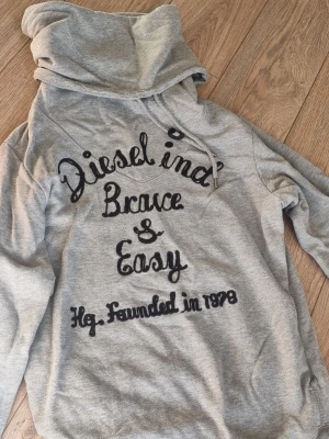 Grå hoodie från Diesel med tryck - Snygg grå hoodie från Diesel med stor krage och dragsko. Framsidan har svart broderat texttryck: 'Diesel ind. Brave & Easy Hg. Founded in 1978'. Tillverkad i mjukt bomullsmaterial och har en avslappnad passform. Perfekt för en chill och trendig look.