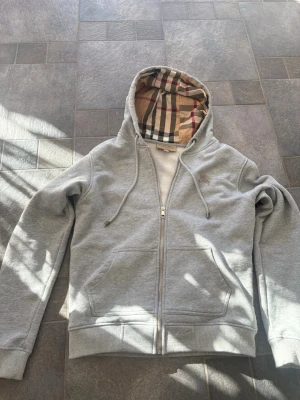 Grå zip hoodie från Burberry - Säljer en grå hoodie från Burberry med dragkedja och klassiskt rutigt foder i huvan. Hoodien har ribbade muddar, fickor framtill och är gjord i mjukt bomullsmaterial. Perfekt för dig som gillar stilrena plagg med en twist från ett ikoniskt märke.