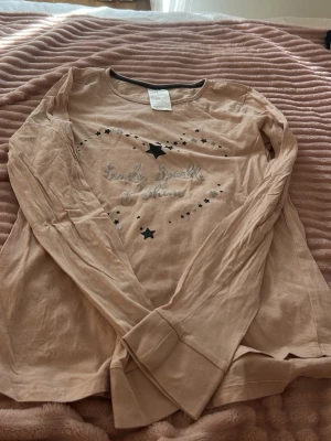 Beige långärmad pyjamastopp med stjärnor - Mysig beige pyjamastopp med långa ärmar och tryck av svarta stjärnor samt texten 'Only sparkle at night' framtill. Rund halsringning med kontrastkant. Perfekt för chilliga kvällar och nätter.