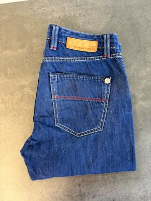 Blå jeans från TRAMAROSSA W32 - Snygga blå jeans från Tramarossa med raka ben och klassisk femficksdesign. Jeansen har kontrastsömmar i vitt och rött samt en läderpatch bak i midjan. Tillverkade i slitstarkt denimtyg, perfekta för en avslappnad och trendig look.