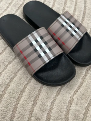 Rutiga slides . Bürbęrr y - Säljer ett par klassiska slides  i storlek 44. De har svart sula och bred rem med det ikoniska beige, svarta, vita och röda rutmönstret. Perfekta för chill dagar eller sommarhäng. Tillverkade i syntetmaterial med bekväm passform.