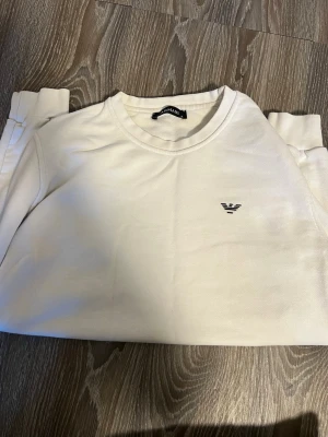 Vit sweatshirt från Armani - Snygg vit sweatshirt från Armani med rund halsringning och diskret logga broderad på bröstet. Tröjan har en clean look och är tillverkad i mjukt material som känns skönt mot huden. Perfekt för en stilren och avslappnad outfit.