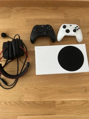 Xbox Series S med två kontroller och headset - Säljer en vit Xbox Series S 512GB med stilren design och svart ventilationsgaller. Medföljer två trådlösa handkontroller, en svart och en vit, samt ett gamingheadset med mikrofon och röda detaljer. Perfekt för gamingkvällar och multiplayer.