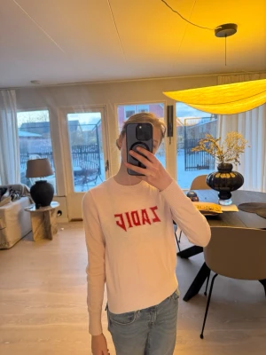 Ljusrosa stickad tröja Zadig & Voltaire - Ljusrosa stickad tröja från Zadig & Voltaire med röd text 'ZADIG' på bröstet. Tröjan har rund halsringning, långa ärmar och ribbade muddar. Perfekt för dig som gillar en clean men ändå statement look.
