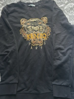 Svart sweatshirt med tiger från Kenzo - Cool svart sweatshirt från Kenzo med ikonisk broderad tiger i gult, beige och brunt på bröstet samt texten 'KENZO PARIS'. Tröjan har rund halsringning och ribbade muddar. Perfekt för dig som vill sticka ut med en statement-piece.