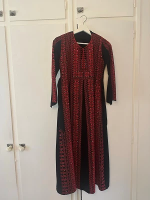 Svart långklänning med rött broderi - Unik svart långklänning med detaljerat rött broderat mönster längs hela klänningen och trekvartsärm. Klänningen har en rak passform och rund halsringning. Perfekt för dig som vill sticka ut med traditionell och färgstark design.
