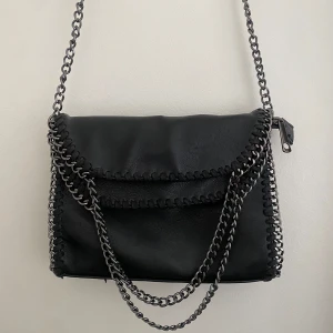 Svart axelväska med kedjedetaljer - Stella McCartney liknande snygg svart axelväska med coola silverfärgade kedjor som axelrem och runt kanterna. Väskan har lockstängning, dekorativa sömmar och en dragkedjeficka på baksidan. Perfekt storlek för det viktigaste och ger en edgy vibe till din outfit. Knappt använd