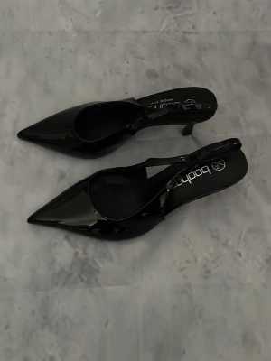 Svarta lackpumps från Boohoo - Snygga svarta pumps i lack från Boohoo med spetsig tå och öppen häl. Skorna har en smal rem runt hälen och en elegant siluett som ger en trendig look. Perfekta för dig som vill sticka ut med stilrena detaljer och glansigt material.