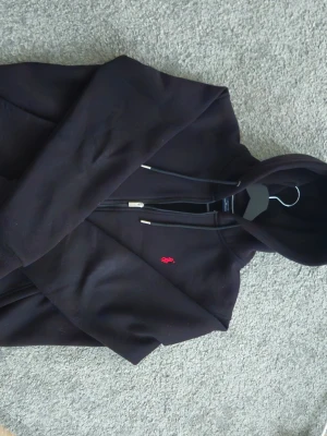 Svart hoodie från Polo Ralph Lauren - Säljer en svart hoodie från Polo Ralph Lauren med dragkedja och huva. Klassisk röd broderad logga på bröstet och justerbara snören i huvan. Mjuk och skön bomullsblandning, perfekt för en avslappnad stil. Inga skador.