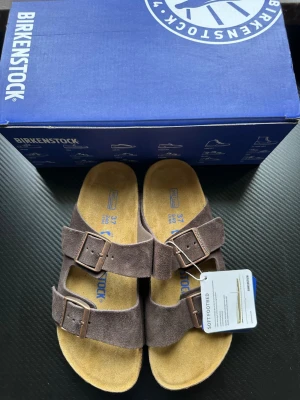 Bruna Birkenstock Arizona sandaler - Birkenstock Arizona sandaler i brun mocka med två breda remmar och justerbara metallspännen. Fotbädden är mjuk och har en naturlig korksula med greppvänlig yttersula. Klassisk och tidlös design, perfekt för dig som gillar komfort och stil.