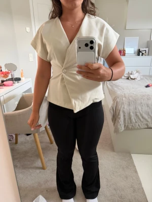 Beige omlottopp med knapp från Sisters Point - Stilren beige omlottopp från Sisters Point med v-ringning och korta ärmar. Toppen har en dekorativ knapp framtill som ger en snygg detalj. Perfekt till både jeans och kjol för en trendig look.