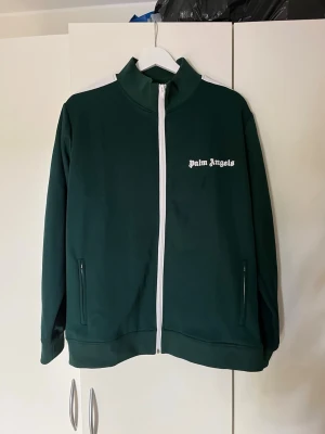 Grön ziptröja från Palm Angels - Mörkgrön ziptröja från Palm Angels med vit dragkedja och vita detaljer på axlarna. Tröjan har hög krage, två fickor med dragkedja och logga i vitt på bröstet. Perfekt för en sportig och trendig look.