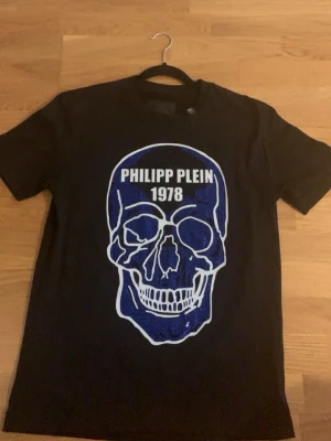 Svart Philipp Plein t-shirt - Snygg svart t-shirt från Philipp Plein med stor blå och vit döskallegrafik och texten 'PHILIPP PLEIN 1978' på bröstet. Klassisk passform och rund hals. Perfekt för dig som gillar statement-plagg och streetwear vibes.