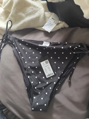Svart prickig bikiniunderdel med knyt - Snygg svart bikiniunderdel från Gina Tricot med vita prickar och knyt i sidorna. Perfekt för stranden eller poolen. Klassisk modell som är lätt att matcha med olika överdelar. Materialet är mjukt och bekvämt syntet.