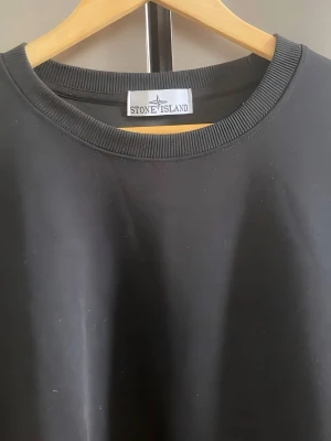 Stone Island tröja  - En stilren svart sweatshirt från Stone Island med klassisk rund halsringning och ribbade muddar. Tröjan har den ikoniska Stone Island-loggan på vänster ärm. Perfekt för dig som gillar streetwear och enkel design.