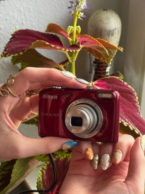 Röd Nikon Coolpix digitalkamera - Snygg röd Nikon Coolpix digitalkamera med kompakt design och silverfärgat objektiv. Kameran har en stor display på baksidan och tydliga knappar för enkel navigering. Perfekt för dig som vill ta skarpa bilder och ha en smidig kamera i fickan. Tillverkad i plast och metall.