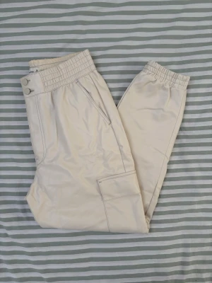 Abercrombie & Fitch krämfärgade byxor i veganskt läder, storlek M - Byxorna har en resårlinning och två knappar framtill. Det finns fickor i sidorna och cargofickor på benen. Bensluten är elastiska. 